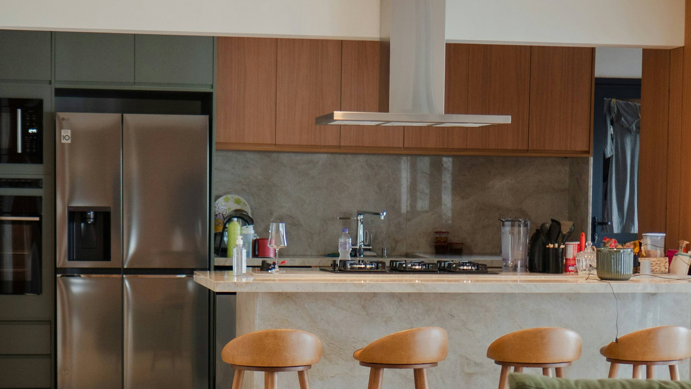 Open-plan kitchen with deep green tall cabinets, walnut upper cabinets, a stainless steel island hood, and a stone waterfall island with four leather-topped bar stools