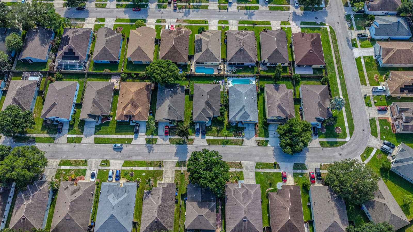 Straight-down aerial view of a Florida single-family subdivision. Rows of detached one-story houses with hip roofs in brown, grey and tan asphalt shingle sit on small rectangular lots along two parallel streets, with a curving connector street and more houses at the right edge. Fenced back yards run between the rows; two hold open swimming pools and others have screened pool enclosures. Concrete driveways with parked cars lead out to sidewalks, and mature shade trees are scattered between the lots.