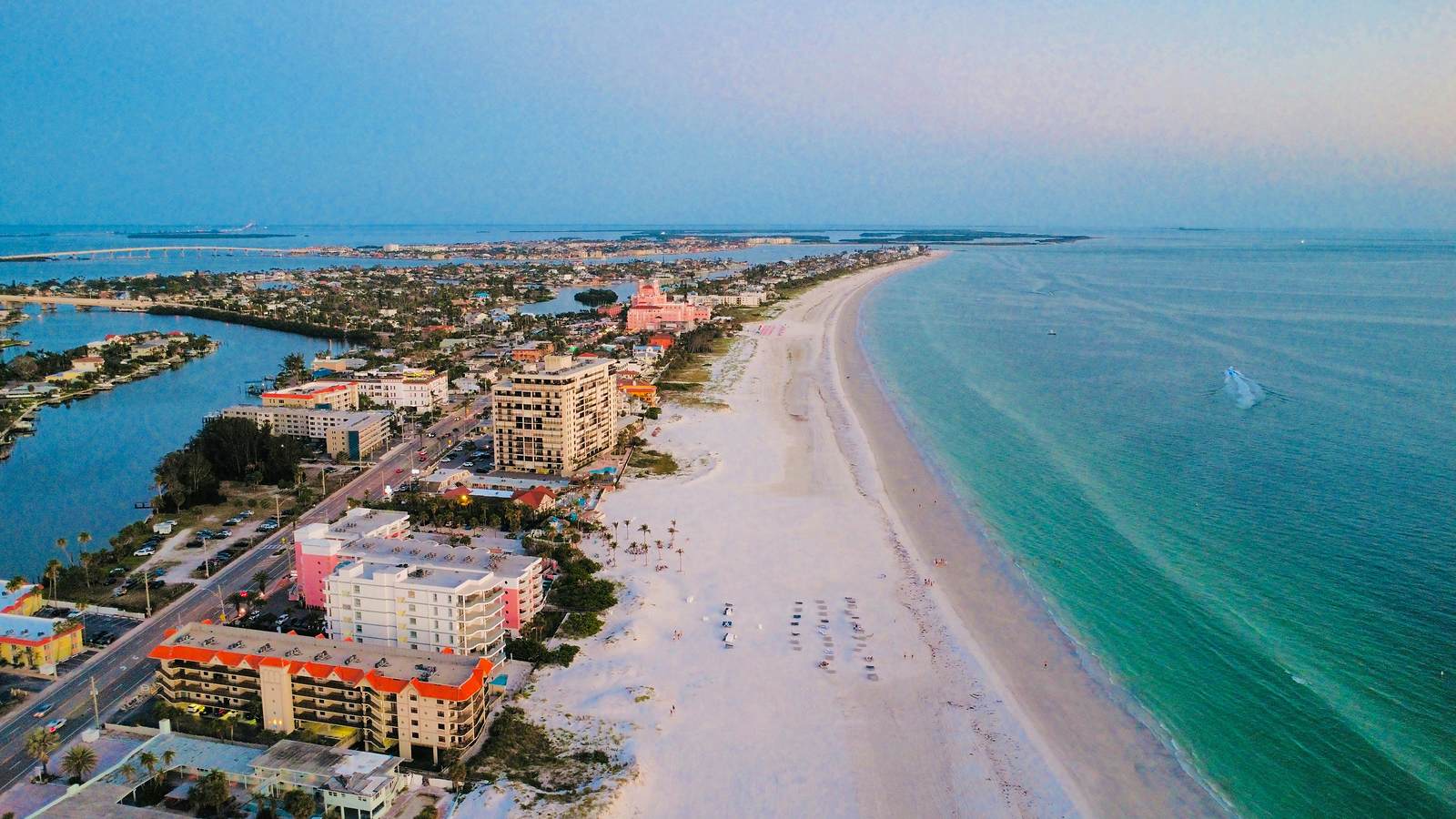 Aerial view along St. Pete Beach at dusk. Low-rise and mid-rise condominium buildings line Gulf Boulevard on the left, a wide white sand beach with rows of beach chairs runs through the center, and the turquoise Gulf of Mexico fills the right side. The pink Don CeSar hotel stands mid-frame, with Boca Ciega Bay and the barrier island stretching to the horizon.