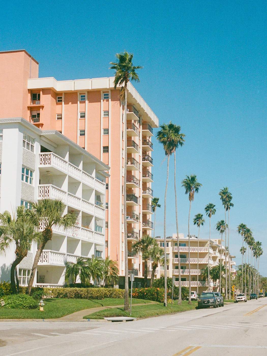 Street-level view of mid-century condominium buildings on a palm-lined St. Petersburg street. In front, a white low-rise shows five tiers of open-air balconies with decorative pierced-concrete balustrades, solid angular concrete privacy dividers between units, and exposed concrete slab edges. Behind it a coral pink tower stacks open balconies with dark metal railings, and a third cream-colored building with white balcony railings continues down the block.