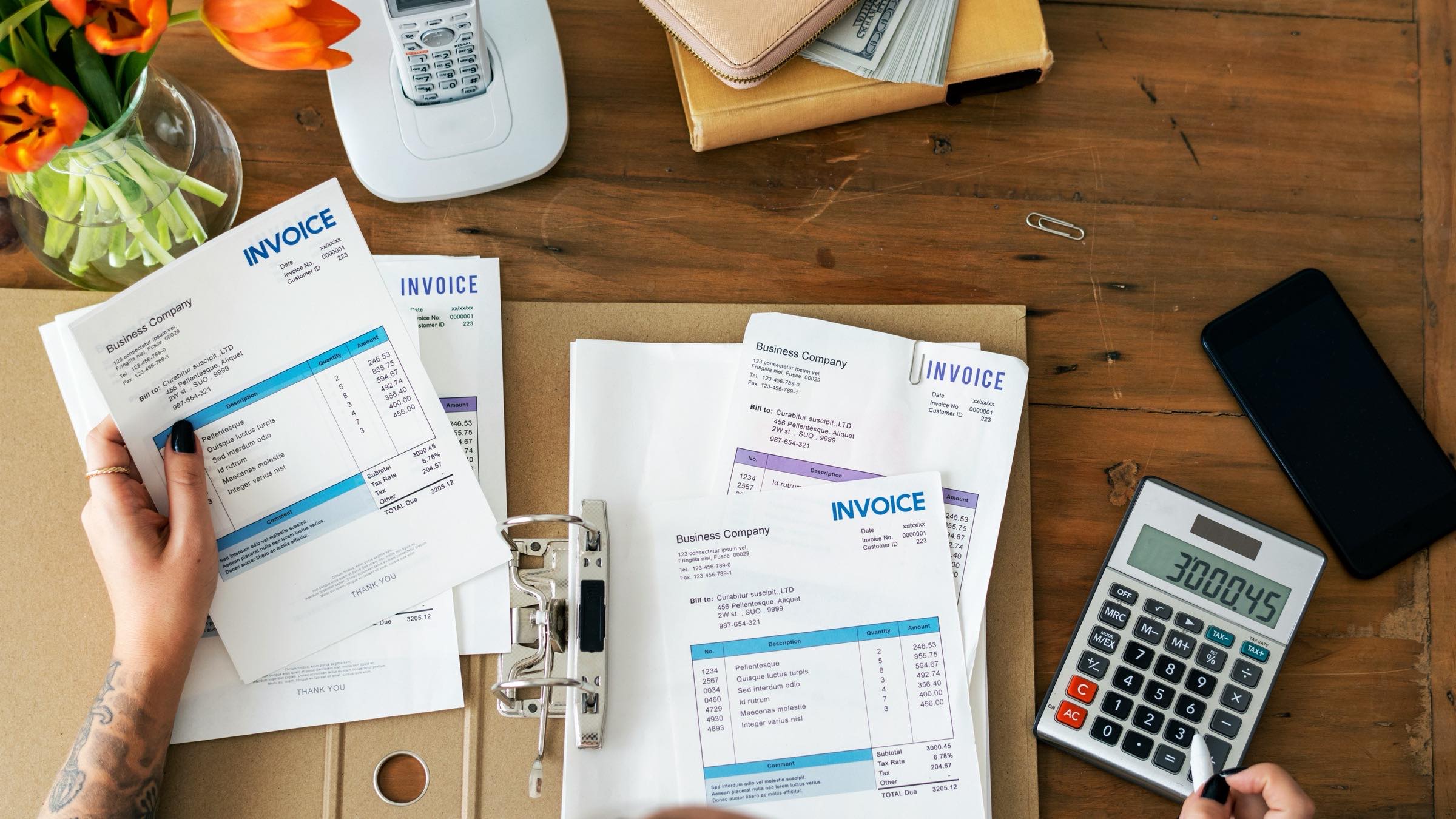 Invoices and receipts spread across a desk beside a binder, a calculator and a phone, with a person reviewing the paperwork