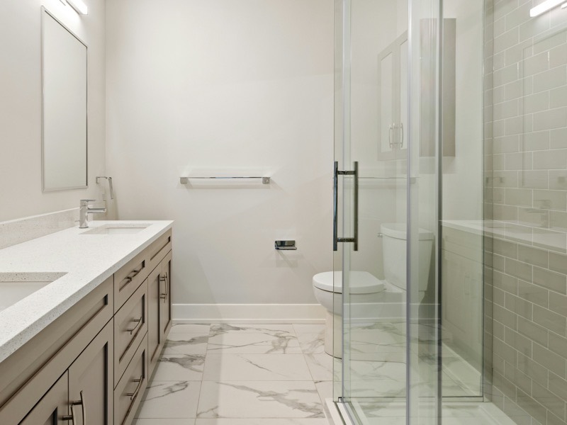 A guest bathroom with a double quartz vanity in taupe shaker cabinets, two mirrors under linear lights, marble-look porcelain floor tile, and a frameless glass shower tiled in pale green subway tile