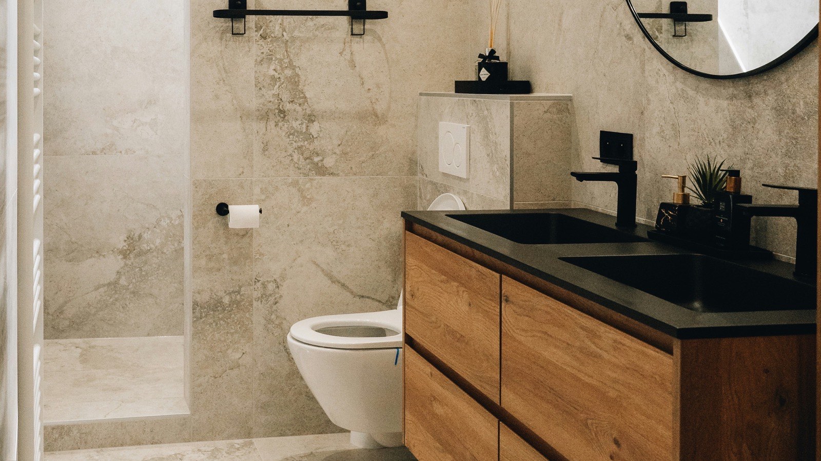 A bathroom finished in large-format stone-look porcelain, with a curbless walk-in shower at left, a wall-hung toilet, a floating oak double vanity with a black stone top, and a round black-framed mirror