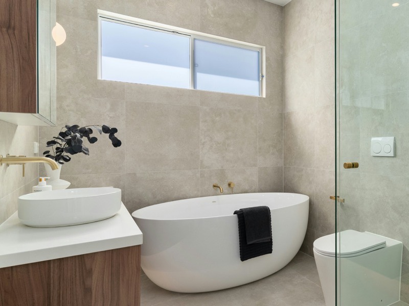 A bathroom with a freestanding oval tub beneath a clerestory window, a frameless glass shower enclosure with a recessed niche, brushed gold fixtures, and a walnut vanity with a round vessel sink