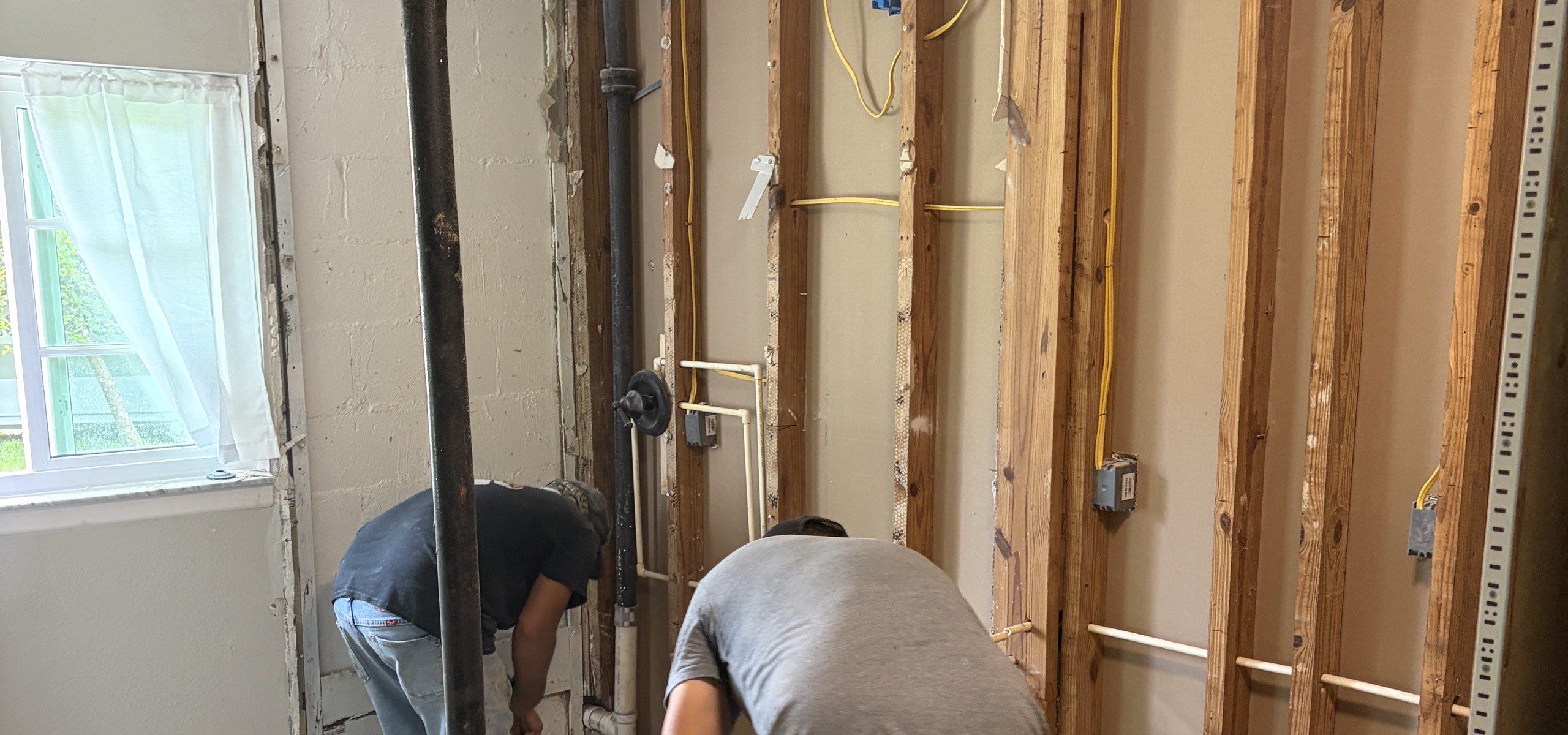 Two workers demolishing a bathroom, one wall opened to bare studs with new plumbing and electrical rough-in running through the bays, a cast iron stack at the centre of the frame