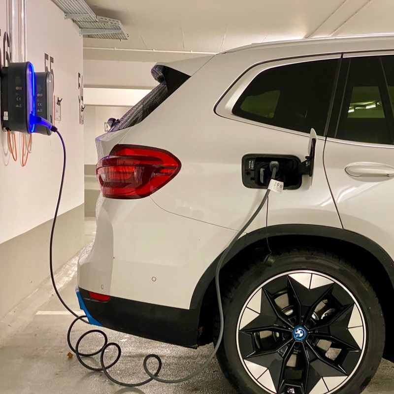 A white electric SUV plugged into a wall-mounted charging unit, the cable running from the charger to the car's charge port