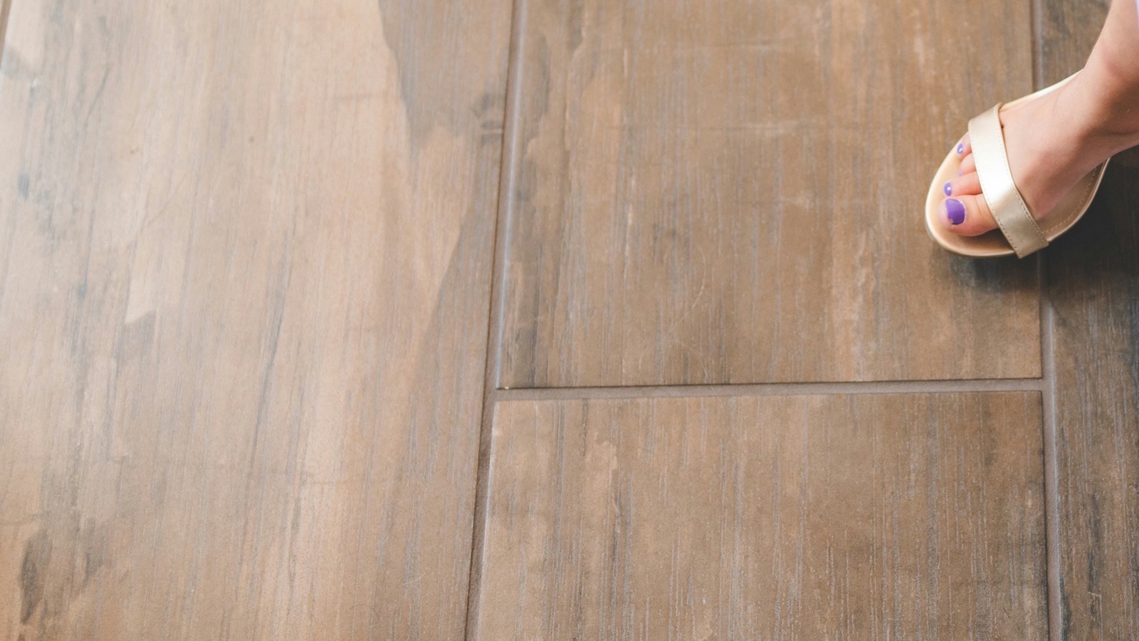 Wood-look porcelain plank tile photographed from above, laid in a staggered pattern with narrow grout joints, a sandal at the top right corner for scale