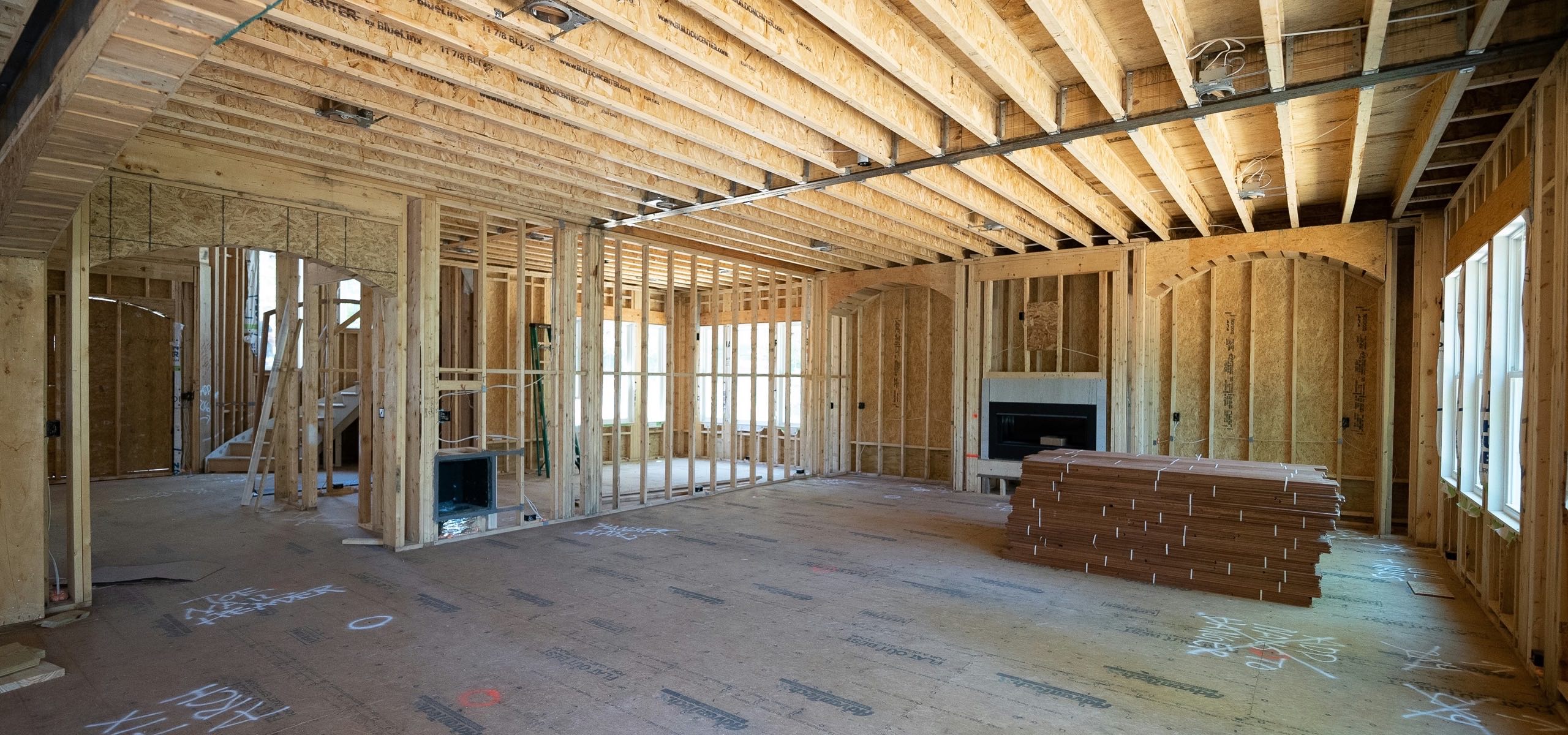 The interior of a house framed out to bare studs, with ceiling joists running overhead, window openings along two walls, a masonry fireplace box, and a stack of lumber on a plywood subfloor covered in chalk layout marks