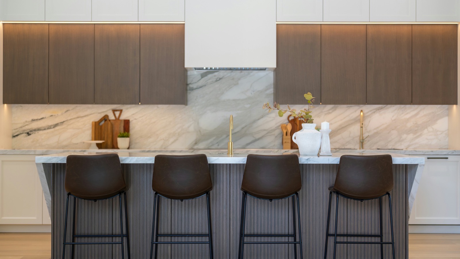 A kitchen elevation with white and walnut upper cabinets flanking a concealed hood, a full-height veined marble slab backsplash, and four leather bar stools at a fluted island