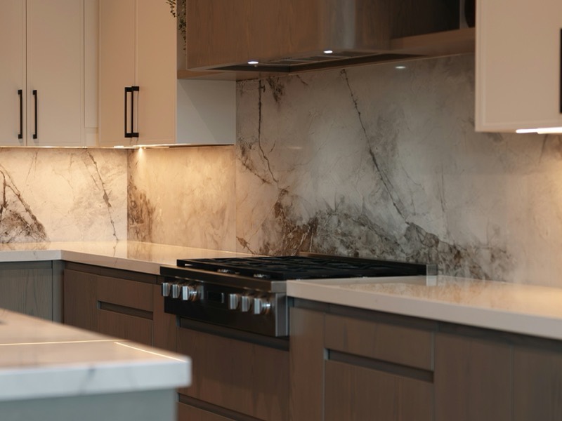 Close view of a kitchen range wall: a gas cooktop under a concealed hood, quartz counters either side, and a heavily veined marble backsplash lit from beneath the upper cabinets