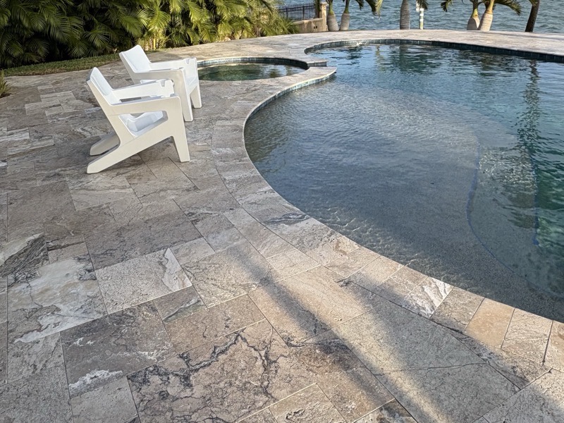 Silver-grey travertine pavers laid in a French pattern around the curved coping of a freeform pool, with two white chairs and a spa spillover at the far edge