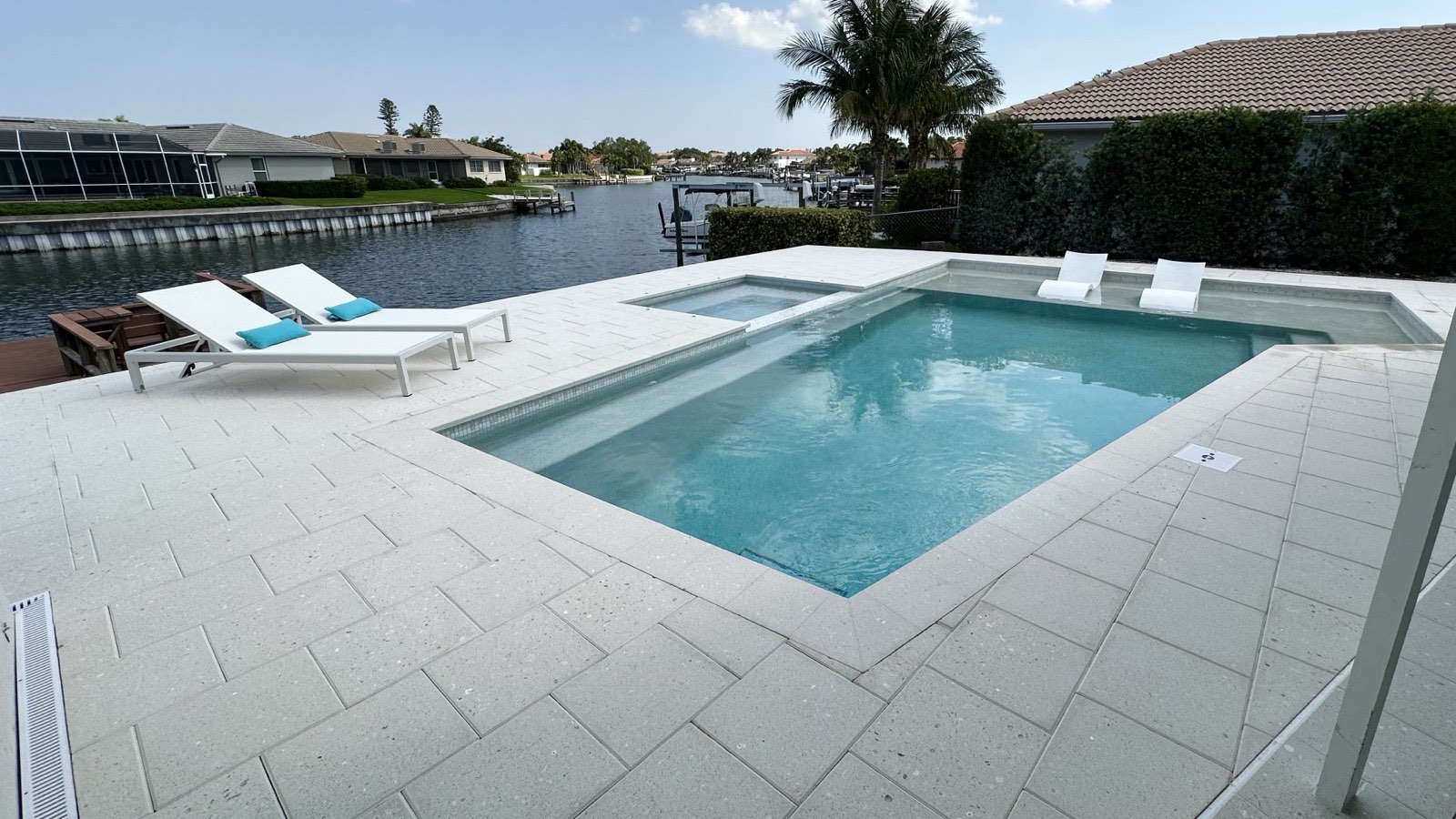A pale paver pool deck on a canal lot, with a rectangular pool and raised spa, two white chaise loungers, and a seawall and neighbouring houses across the water